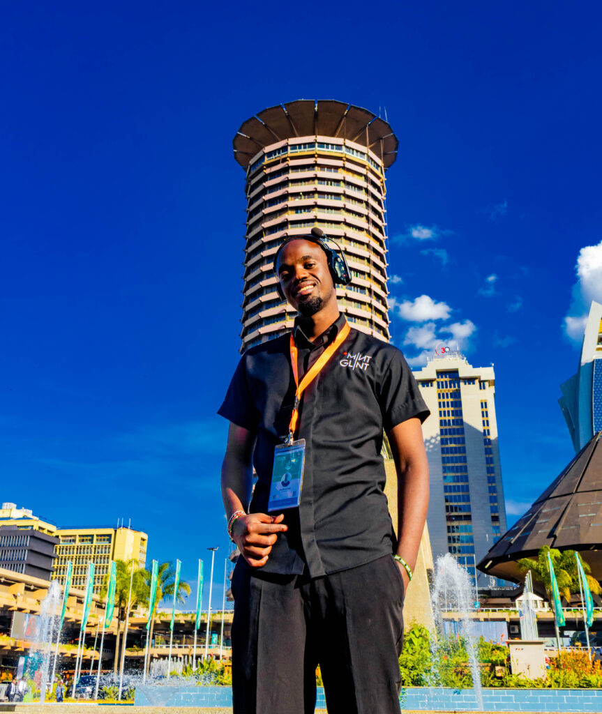 A standing portrait of Kelvin Masila looking confidently over his shoulder directly at the camera. He is in a warmly lit, busy event production area, wearing a blue event crew vest and an orange lanyard over a patterned shirt.