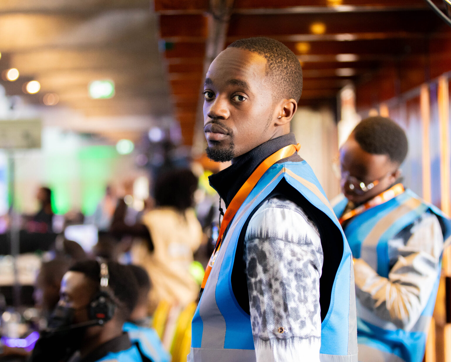 Kelvin Masila sitting at a production desk wearing a blue safety vest and a heavy-duty communications headset. He is looking down attentively at his workstation, with a smartphone resting on the desk beside him.